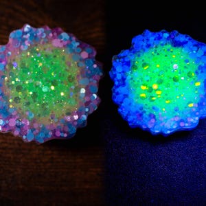 May include: Two resin art pieces with a floral shape. One is a mix of green, pink, and blue glitter. The other glows with blue and green hues. The pieces are on a dark surface.