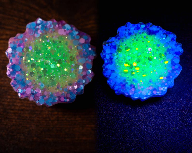 May include: Two resin art pieces with a floral shape. One is a mix of green, pink, and blue glitter. The other glows with blue and green hues. The pieces are on a dark surface.