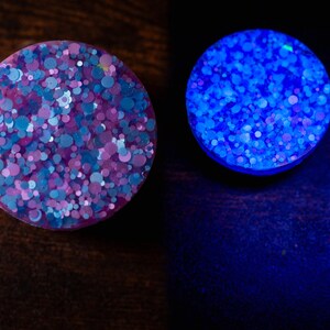 May include: Two round, decorative plugs. One plug is a mix of blue, purple, and pink glitter. The other plug glows bright blue. The plugs are on a dark brown surface.