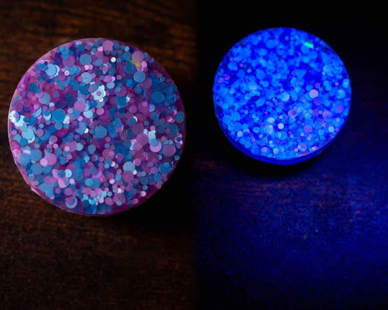 May include: Two round, decorative plugs. One plug is a mix of blue, purple, and pink glitter. The other plug glows bright blue. The plugs are on a dark brown surface.