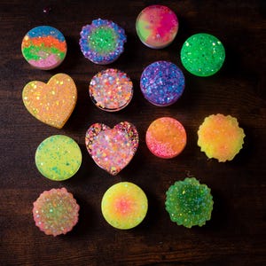 May include: A collection of colorful, glittery phone grips in various shapes, including circles and hearts. The grips feature a range of colors, such as pink, green, orange, and purple, with a sparkling, textured appearance. The items are arranged on a dark brown wooden surface.