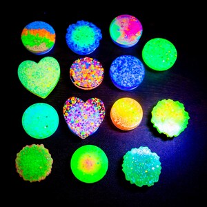 May include: A collection of colorful, glowing accessories in various shapes, including circles and hearts. The accessories feature vibrant colors like green, blue, orange, and pink, with glitter and textured surfaces. The items are arranged on a dark background.