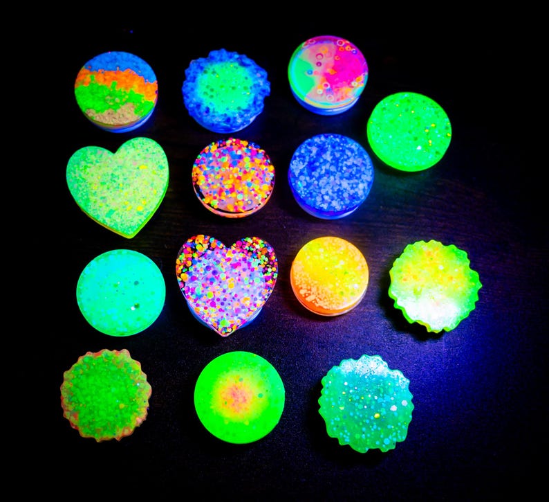 May include: A collection of colorful, glowing accessories in various shapes, including circles and hearts. The accessories feature vibrant colors like green, blue, orange, and pink, with glitter and textured surfaces. The items are arranged on a dark background.