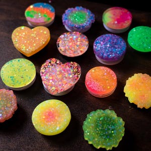 May include: A collection of colorful, glitter-covered phone grips in various shapes, including round and heart-shaped designs. The grips feature a range of colors, such as orange, green, blue, and pink, with a sparkling finish. The items are arranged on a dark surface.