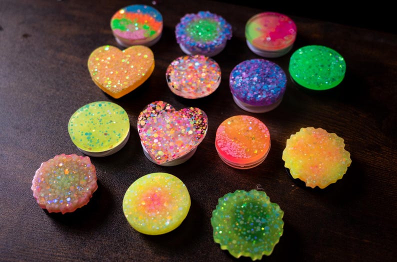 May include: A collection of colorful, glitter-covered phone grips in various shapes, including round and heart-shaped designs. The grips feature a range of colors, such as orange, green, blue, and pink, with a sparkling finish. The items are arranged on a dark surface.