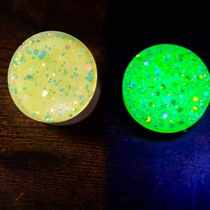 May include: Two round, glitter-filled plugs. One is a pale yellow with green and pink glitter. The other glows bright green with colorful glitter. The plugs are likely for ear piercings.
