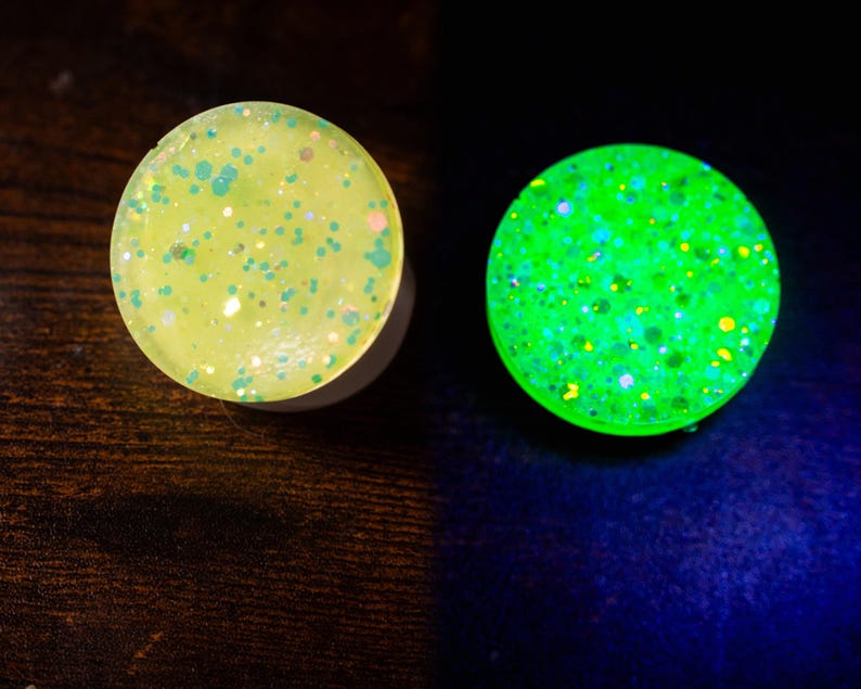 May include: Two round, glitter-filled plugs. One is a pale yellow with green and pink glitter. The other glows bright green with colorful glitter. The plugs are likely for ear piercings.