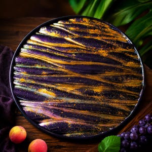 May include: A round serving tray with a black rim. The tray has a textured, wave-like pattern in deep purple and gold. Two peaches and a bunch of grapes are visible, suggesting a food presentation theme. The tray's design is artistic and decorative.