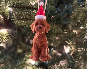 Könnte beinhalten: Ein braunes, lockiges Hundeornament mit strukturiertem Fell, schwarzen Augen und einer schwarzen Nase. Der Hund trägt eine kleine, leuchtend rote Weihnachtsmütze mit weißem Rand und weißem Bommel, aufgehängt an einer dünnen weißen Schnur.