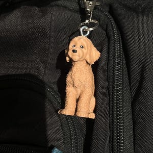 Può includere: Un ciondolo a forma di cane barboncino color marrone chiaro, attaccato a un moschettone in metallo argentato. Il ciondolo presenta un aspetto di pelliccia strutturata e pende su uno sfondo nero.