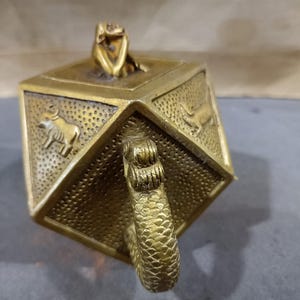 Puede incluir: Un recipiente de latón antiguo detallado, con forma geométrica. Presenta figuras de animales en relieve y un asa con textura escamosa. Una pequeña figura de animal está sobre la tapa. El diseño sugiere un artículo vintage o de colección.