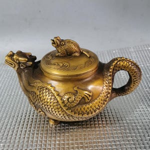 Puede incluir: Una tetera de bronce detallada con motivos de dragones y ranas. La tetera presenta una boquilla con cabeza de dragón y un cuerpo con escamas de dragón. Una rana se sienta sobre la tapa. El asa es una cola de dragón.