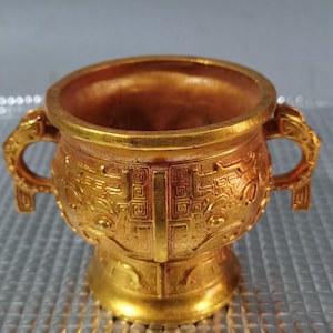 May include: A gold-colored, ornate vessel with two dragon-shaped handles. The vessel features detailed relief patterns and a pedestal base. The interior is a darker gold, contrasting with the exterior's brighter finish. The overall design suggests a ceremonial or decorative object.