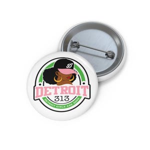 May include: A round pin-back button featuring a graphic of a person wearing a black and pink baseball cap with large gold hoop earrings. The design includes the text DETROIT 313 and DETROIT GIRLS ARE DOPE in pink and green lettering.