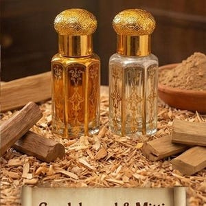Puede incluir: Dos frascos de perfume ornamentados con tapas y detalles dorados, uno lleno de l&iacute;quido &aacute;mbar, el otro transparente. Los frascos est&aacute;n sobre virutas de madera con trozos de s&aacute;ndalo y un cuenco con polvo. Texto: "S&aacute;ndalo y Mitti: Cremoso, riqueza amaderada con el aroma de tierra horneada."