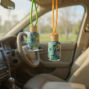 Puede incluir: Dos ambientadores colgantes para coche con estampados florales en tonos azules y verdes. Los ambientadores tienen tapas de madera y están suspendidos por cordones verdes y naranjas. El interior de un coche es visible en el fondo.
