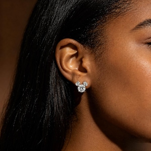 May include: A close-up of a person's ear adorned with a pair of diamond stud earrings. The earrings are designed in a Mickey Mouse silhouette, with a large round center stone and two smaller round stones above.