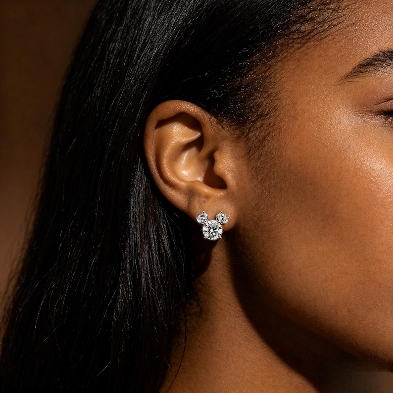 May include: A close-up of a person's ear adorned with a pair of diamond stud earrings. The earrings are designed in a Mickey Mouse silhouette, with a large round center stone and two smaller round stones above.