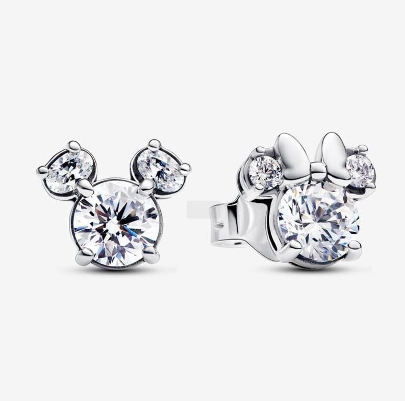 May include: A pair of silver stud earrings. One earring features a Mickey Mouse design with a large, round, clear crystal center and two smaller, pear-shaped crystals. The other earring has a Minnie Mouse design with a bow.