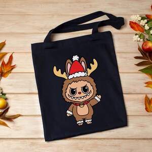 May include: A navy blue tote bag featuring a cartoon character dressed as a reindeer with a Santa hat. The character has large eyes, a toothy grin, and is wearing a red collar with a gold bell. The bag has long black handles.