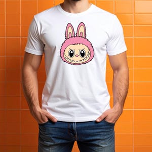 May include: White t-shirt featuring a cartoon illustration of a pink bunny-like creature with a mischievous expression. The creature has large eyes, a toothy grin, and fluffy ears. The shirt is made of a soft material.