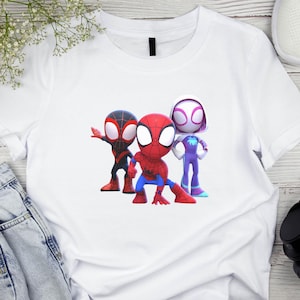 May include: White t-shirt with a cartoon graphic of three Spider-Man characters. The characters are in a cartoon style, with one in red and blue, one in black and red, and one in purple and white. The shirt is made of a soft material.