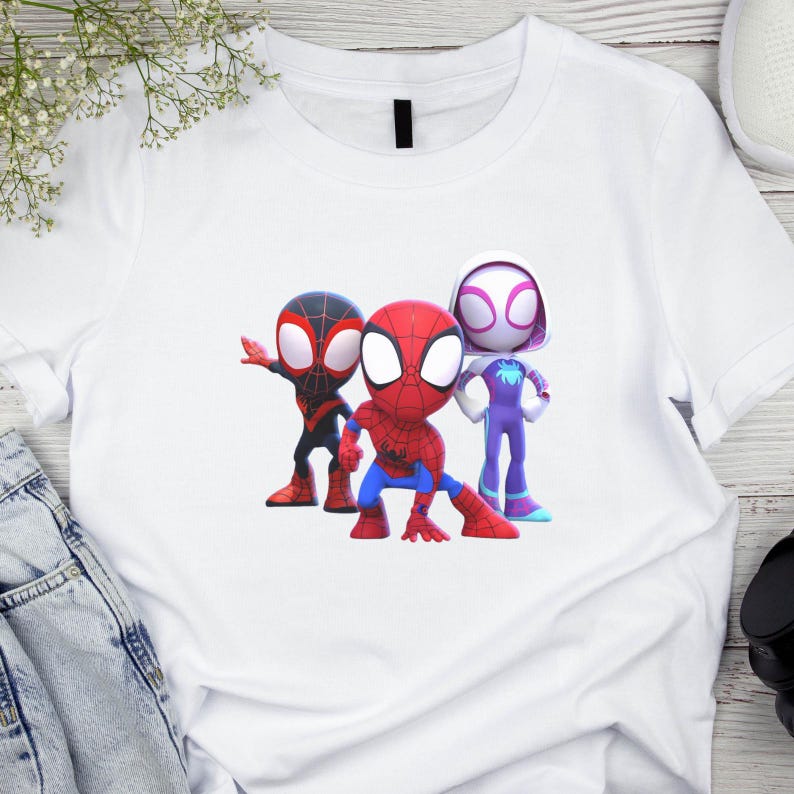May include: White t-shirt with a cartoon graphic of three Spider-Man characters. The characters are in a cartoon style, with one in red and blue, one in black and red, and one in purple and white. The shirt is made of a soft material.