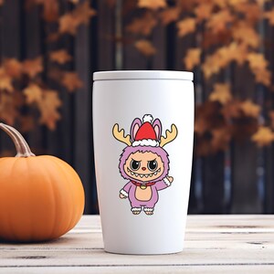 May include: White travel mug featuring a cartoon monster in a purple costume with antlers and a Santa hat. The monster has a red collar and is waving. An orange pumpkin sits to the left of the mug.