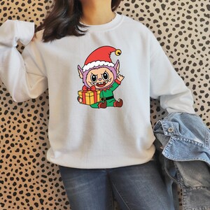 May include: White sweatshirt featuring a cartoon elf wearing a red hat with a gold bell. The elf is holding a yellow gift box with a red bow. The elf is wearing a green outfit with red shoes.