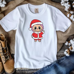 May include: White t-shirt featuring a cartoon character dressed in a Santa Claus outfit. The character has a red Santa hat, a red coat with white trim, and a smiling face. The shirt is on a wooden surface with jeans and boots.