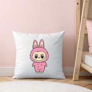 May include: White square pillow featuring a cartoon illustration of a pink monster in a bunny costume. The monster has large eyes, a toothy grin, and is wearing a pink hooded jumpsuit. The pillow is on a light pink background.