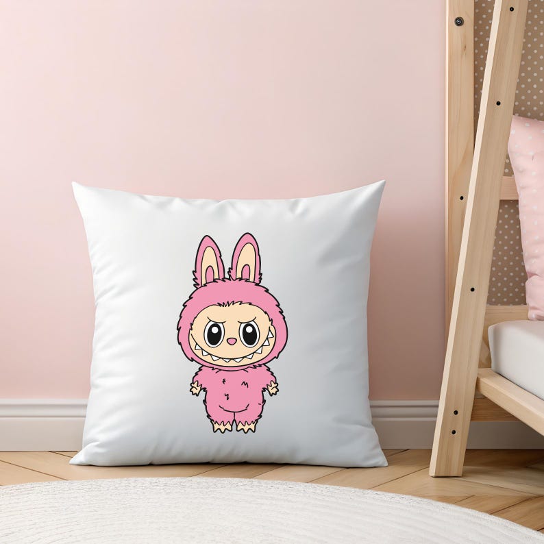 May include: White square pillow featuring a cartoon illustration of a pink monster in a bunny costume. The monster has large eyes, a toothy grin, and is wearing a pink hooded jumpsuit. The pillow is on a light pink background.