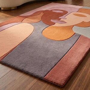 May include: A rectangular area rug with an abstract face design in shades of gray, orange, and pink. The rug is placed on a hardwood floor, with a cream-colored sofa and wooden side table in the background.