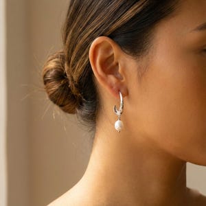 以下が含まれることがあります： テクスチャードデザインと白いパールが揺れるシルバーフープピアス。これらのイヤリングは、普段使いにも特別な機会にも適したクラシックなアクセサリーです。パールがデザインにエレガントさを添えています。