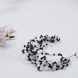 May include: A delicate, handcrafted necklace featuring small, black beads strung on a silver-colored chain. The necklace is arranged in a flowing, layered design. Soft pink flowers are visible in the background, adding a touch of color.