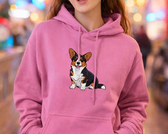 以下が含まれることがあります： コーギー犬のグラフィックが描かれたピンク色のパーカー。コーギーは黒、白、茶色で描かれています。パーカーにはフロントポケットとドローストリングフードが付いています。笑顔の人。