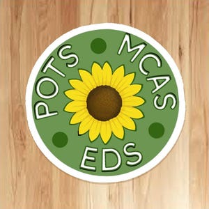 May include: A circular sticker with a green background and a white border. It features a yellow sunflower at the centre and the white text "POTS MCAS EDS" around the flower. There are three dark green dots on the background.