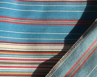May include: A woven fabric featuring a repeating pattern of horizontal stripes in shades of teal, cream, red, yellow, and black. The textile has a visible weave texture and a folded edge showing the reverse side with a similar striped design.