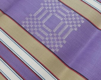 May include: A woven fabric featuring wide vertical stripes in purple and tan, accented by thinner white and blue pinstripes. A repeating geometric square pattern, composed of small white woven squares, is centered within the purple sections.