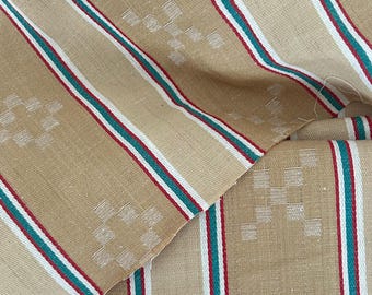 May include: A tan fabric featuring vertical stripes in white, red, and teal. The material displays a repeating geometric pattern of small, light-colored squares arranged in a cross-like shape between the vertical striped sections.
