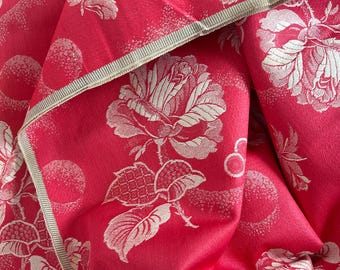 May include: A vibrant hot pink fabric featuring a woven white floral pattern of large blooming roses and circular motifs. The textile has a folded edge finished with a light beige ribbed trim, showcasing a detailed jacquard texture throughout.