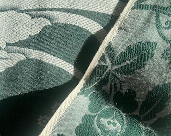 May include: A dark green fabric featuring a woven floral pattern in a lighter shade of green. The textile displays a dense, intricate botanical design with visible leaves and blossoms, finished with a light-colored, woven edge trim.
