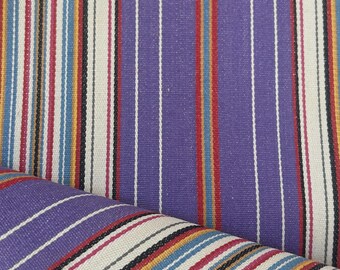 May include: A woven fabric featuring a repeating vertical striped pattern in shades of purple, cream, red, yellow, and blue. The texture shows a tight, durable weave with thin white and black pinstripes separating the wider colored bands.