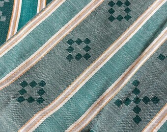 May include: A teal fabric featuring a woven pattern of vertical stripes in white and tan, interspersed with rows of small, dark teal diamond-shaped motifs. The textile has a visible weave texture and a consistent color palette throughout.
