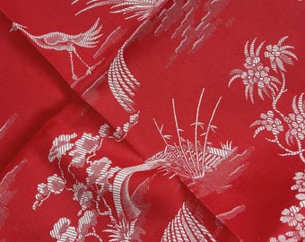 May include: A vibrant red fabric featuring a repeating white pattern of cranes, bamboo stalks, and floral elements. The design is rendered in a fine, detailed line work that contrasts sharply against the solid red background of the textile material.