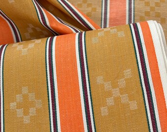 May include: A folded textile featuring vertical stripes in shades of mustard yellow and bright orange, accented by thin white and dark maroon pinstripes. The fabric displays a repeating pattern of small, light-colored geometric diamond shapes.