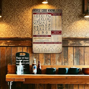 May include: A vintage-style metal sign with Morse code information hangs on a wooden wall. The sign features the alphabet, numbers, and timing instructions. Below, a wooden shelf holds mugs, bottles, and a small chalkboard sign.