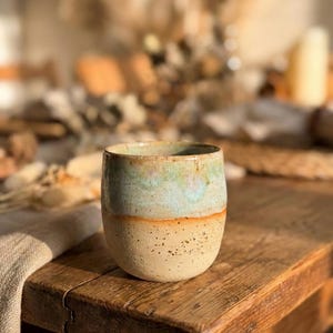 Könnte beinhalten: Eine handgefertigte Keramiktasse mit einem gesprenkelten, beigefarbenen Boden und einem oberen Bereich mit einer Mischung aus Blau-, Grün- und Orangetönen. Die Tasse steht auf einem rustikalen Holztisch.