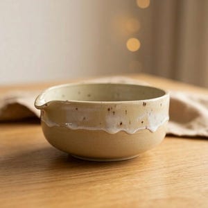 Könnte beinhalten: Eine handgefertigte Keramik-Rührschüssel mit Ausguss. Die Schüssel ist beige mit einem weißen, wellenförmigen Rand und gesprenkelten Details. Sie steht auf einer Holzoberfläche, was auf eine Küche oder einen Essbereich hindeutet.