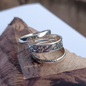 May include: Three silver rings are displayed on a wooden surface. The middle ring features a floral design, while the other two are plain bands. The rings are likely made of sterling silver and are suitable for everyday wear.